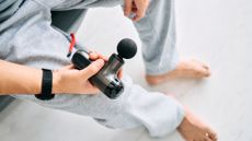 A view looking down on a man wearing gray sweatpants holding a black massage gun in his right hand. 