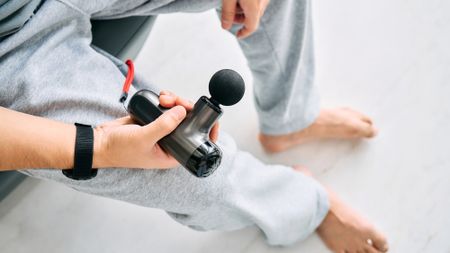 A view looking down on a man wearing gray sweatpants holding a black massage gun in his right hand.