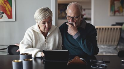 An older couple sit at their dining room table together and look at a laptop screen.
