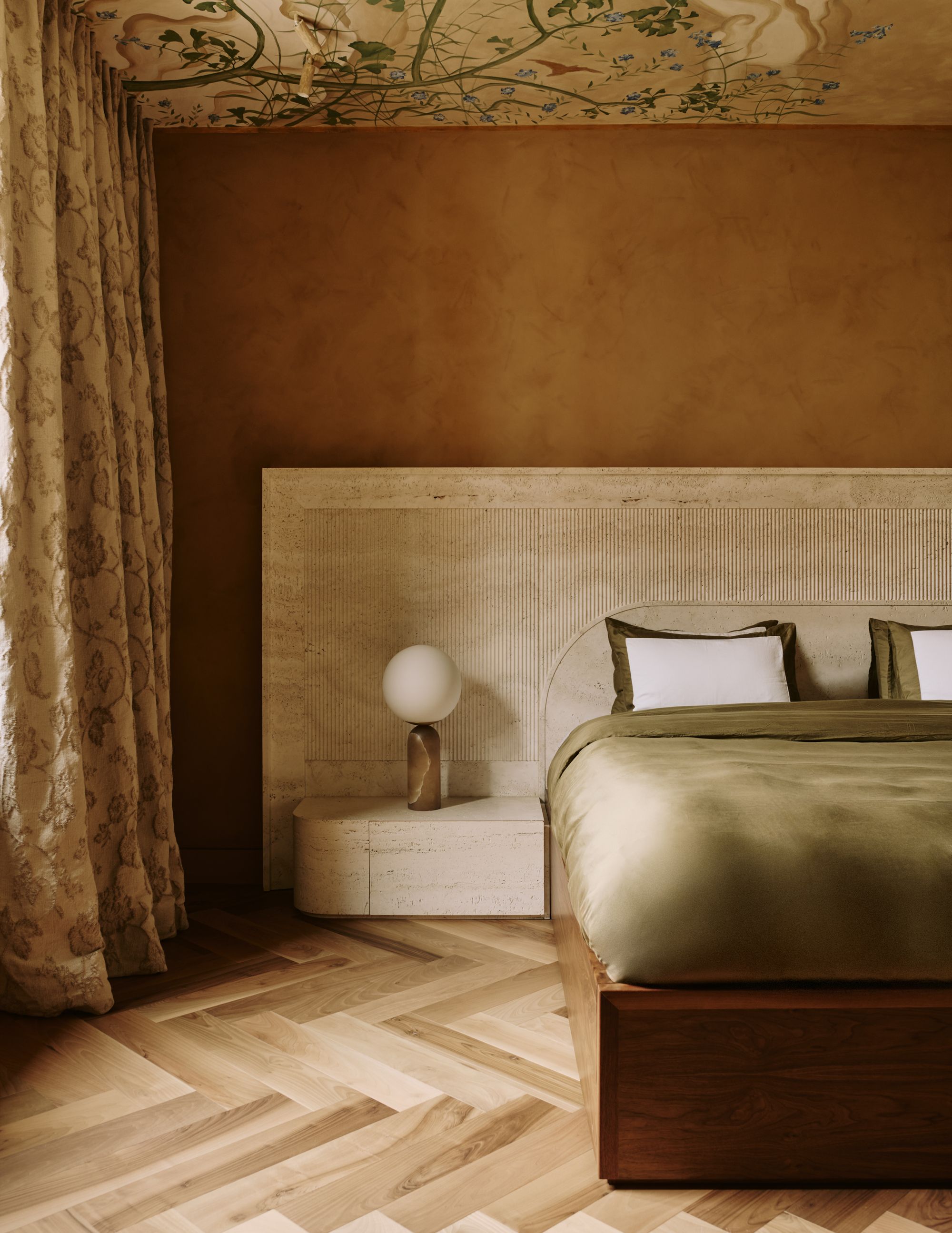 A warm bedroom with burnt orange walls, a bed with a white extended headboard and olive green bedding. The ceiling is painted, and there are patterned curtains on the windows.