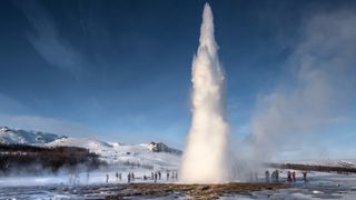 iceland geyser erupting high into the blue sky with people watching below.