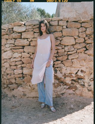 a model stands in front of a stone wall wearing a sheer slip dress over loose pants and metallic sandals