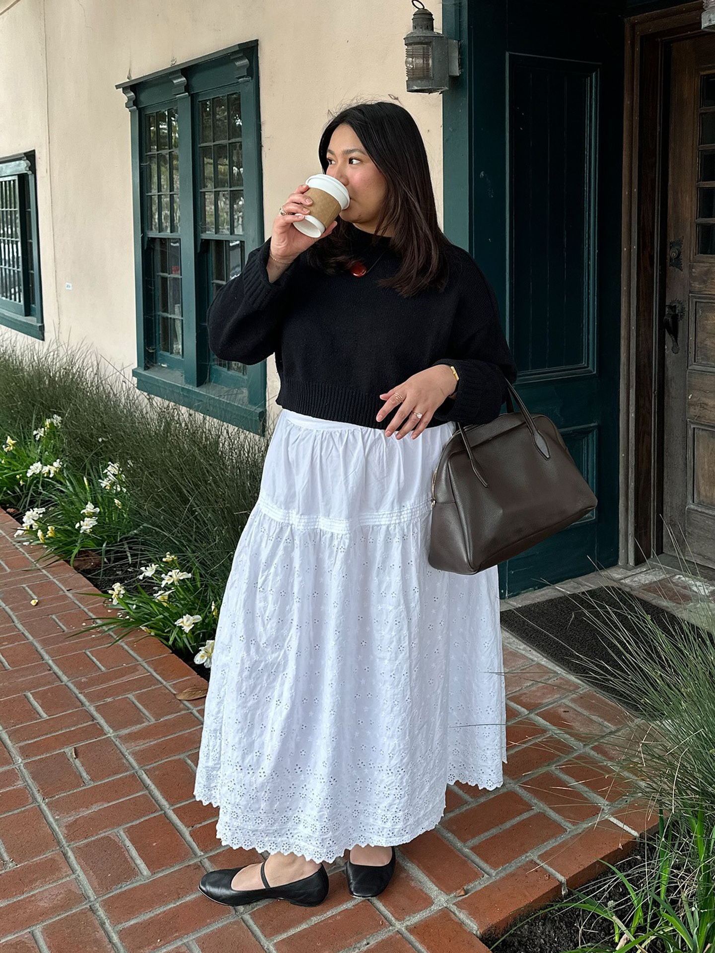 style influencer Marina Torres poses on a brick pathway surrounded by grass and flowers drinking a coffee while wearing a black crewneck sweater, cord necklace with a red pendant, simple brown leather bag, a white tiered poplin skirt and black Mary Jane flats