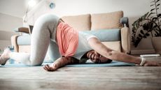 Woman performing thread the needle stretch on the floor with a couch behind her. She is on her knees with one hand on the floor in front of her. The other hand is on the floor to the side between her other arm and knee. She is smiling. 