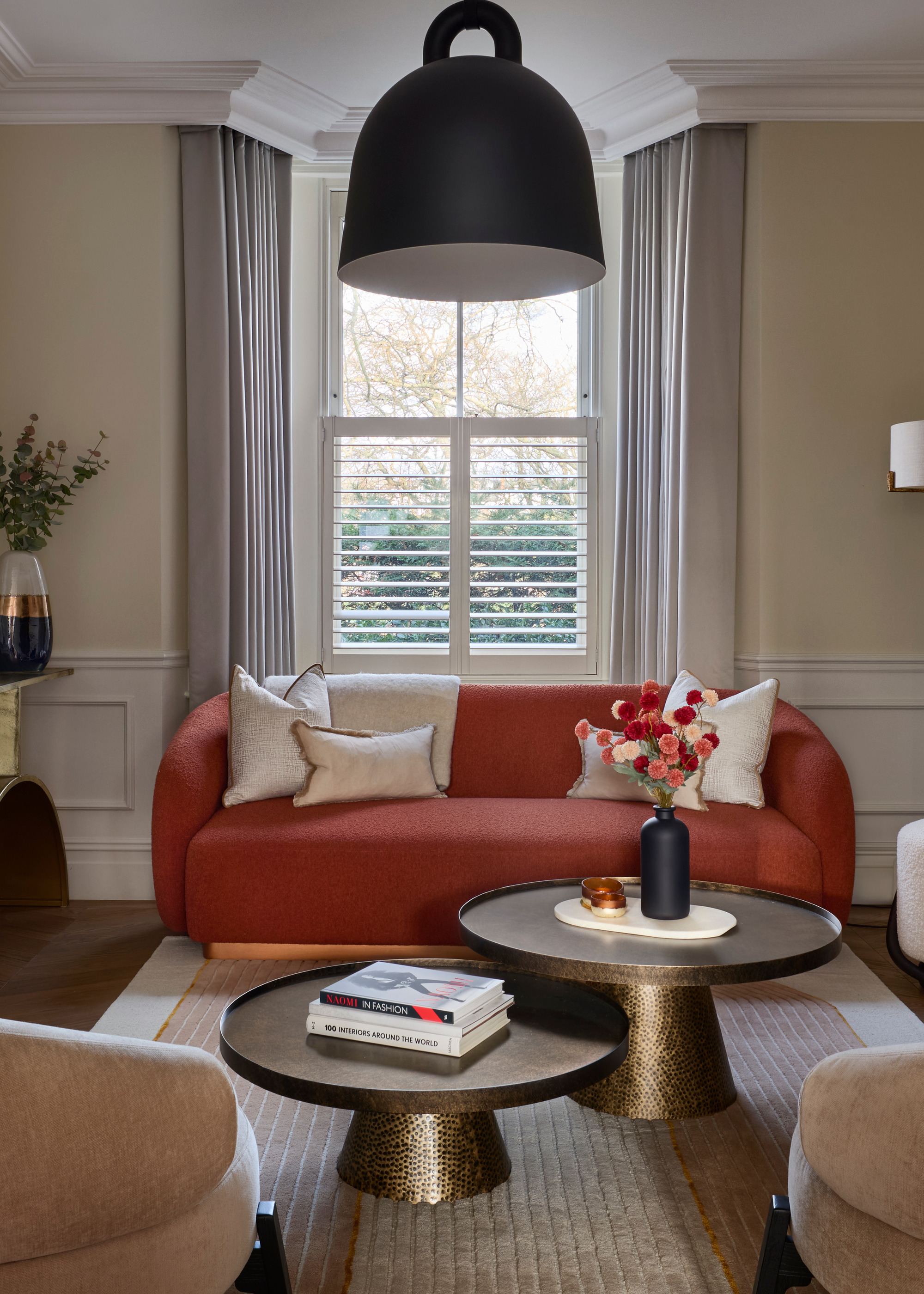 A living room with concealed curtain tracks, bronze side tables, a burnt orange sofa, two curved beige armchairs, and two nesting round bronze tables