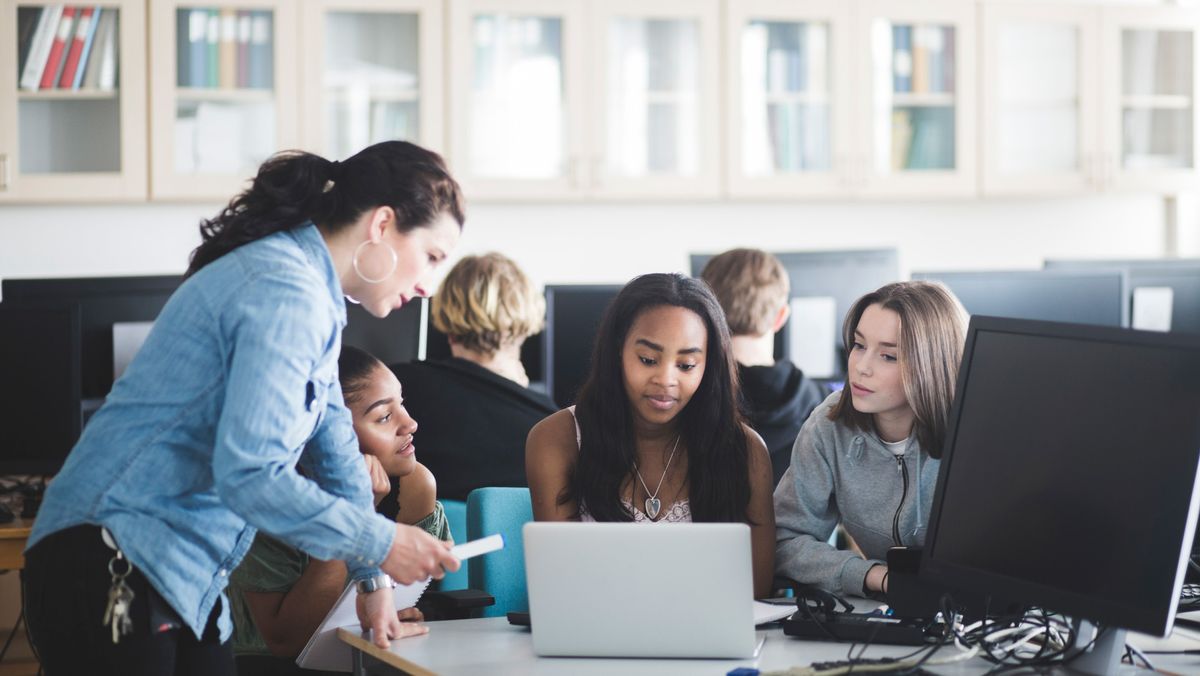 A female teacher explaining something to a small group of female students, who are sat around a desk looking at a laptop. All are sat in their school&#039;s computer lab.