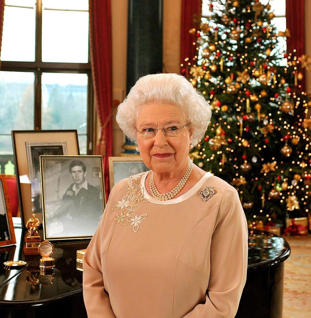 Queen Elizabeth in a tan dress standing in front of a piano and christmas tree