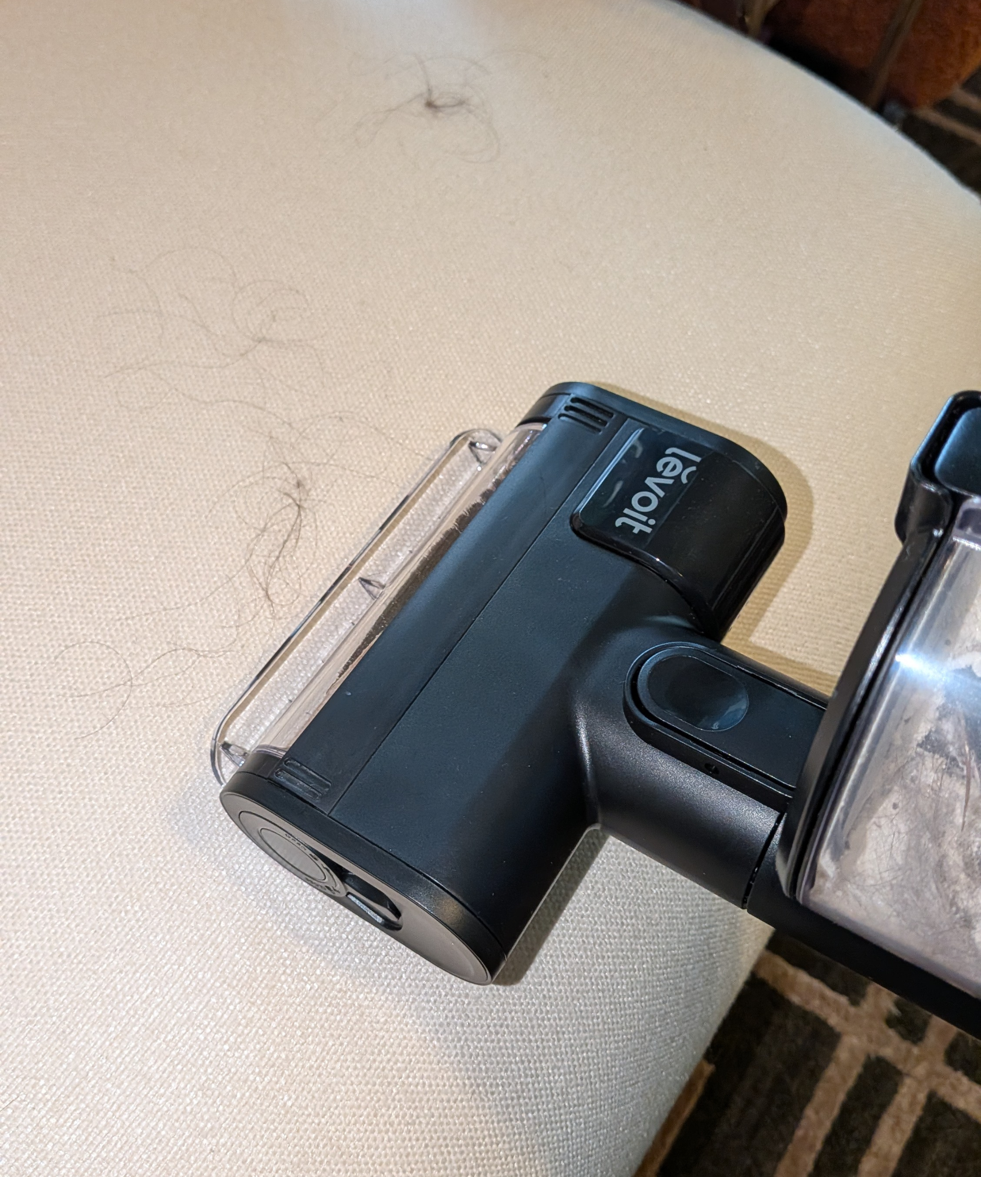 A mini brush has been added to the Levoit VortexIQ Pro vacuum and is being used to suction up hair from a cream-colored armchair.