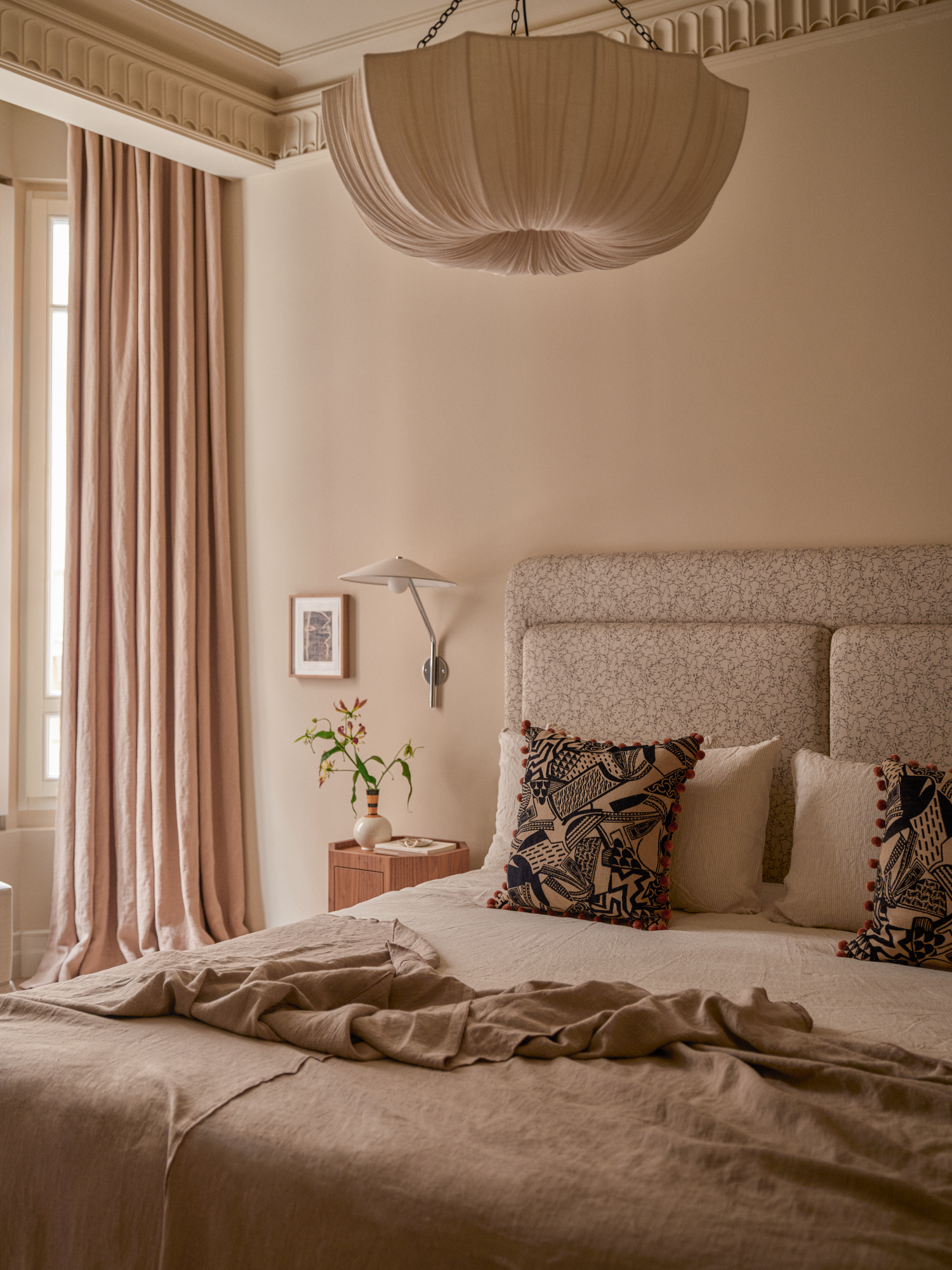 Off-white bedroom with warm grey patterned headboard, pleated ceiling light and soft pink curtains