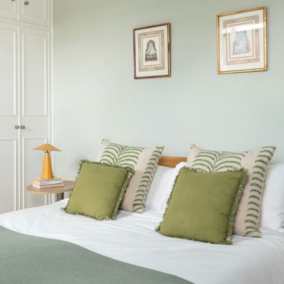 A soft green-painted bedroom with green cushions and throws on the bed and a yellow glossy metal table lamp from H&M on the bedside table 