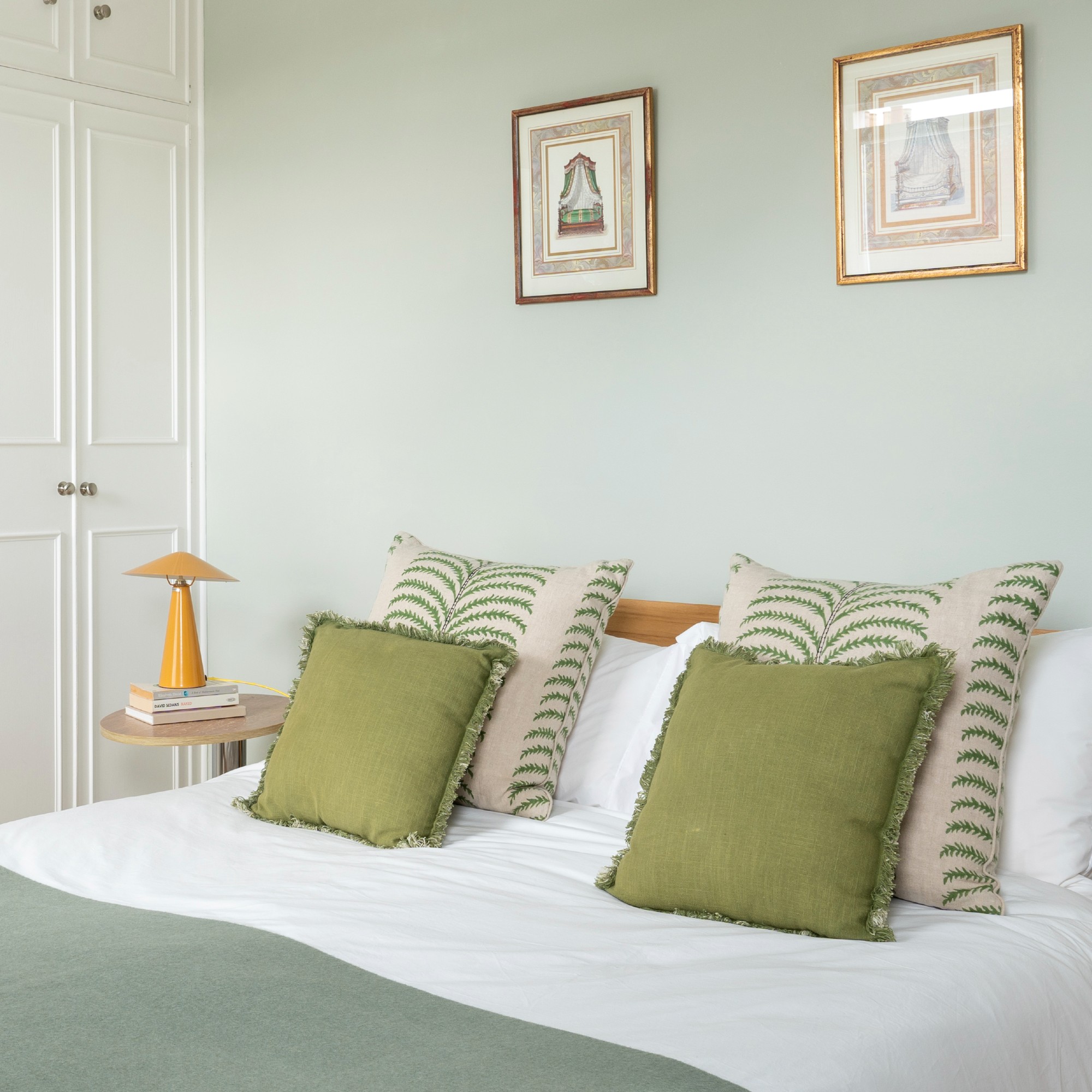 A soft green-painted bedroom with green cushions and throws on the bed and a yellow glossy metal table lamp from H&amp;M on the bedside table 