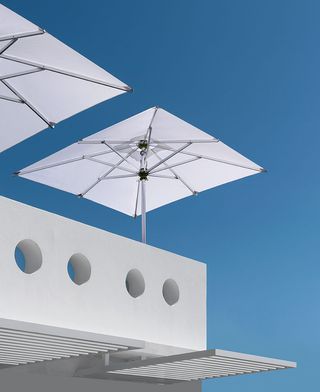 Two white parasols on a balcony with round holes along its sides, white awnings below it and the blue sky behind it.