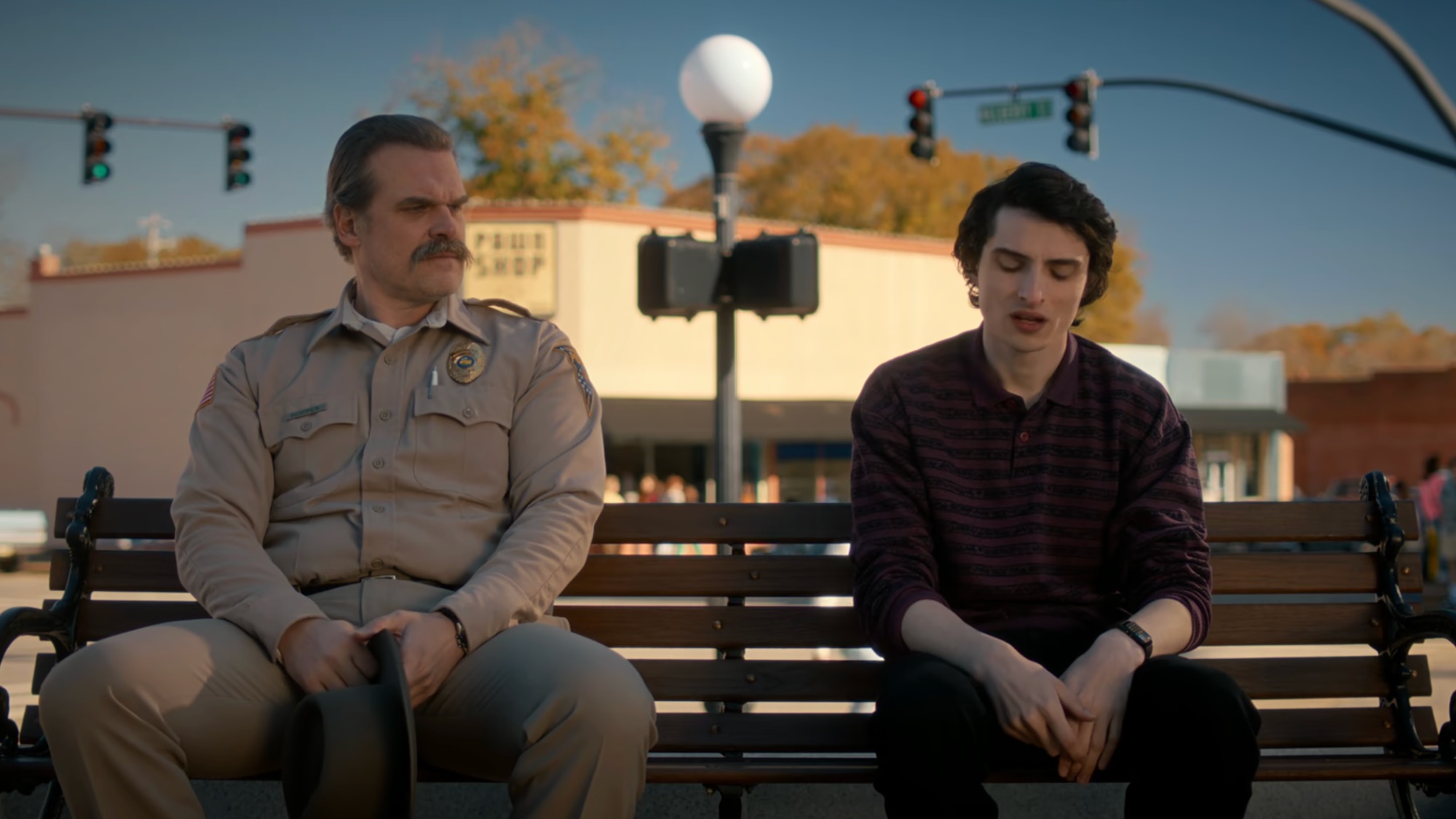 Hopper in his sheriff uniform sitting on a bench next to a somber Mike Wheeler in Downtown Hawkins in Stranger Things Season 5, Episode 8. 