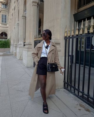 Aida wears a trench coat, white shirt, black tailored shorts and sandals