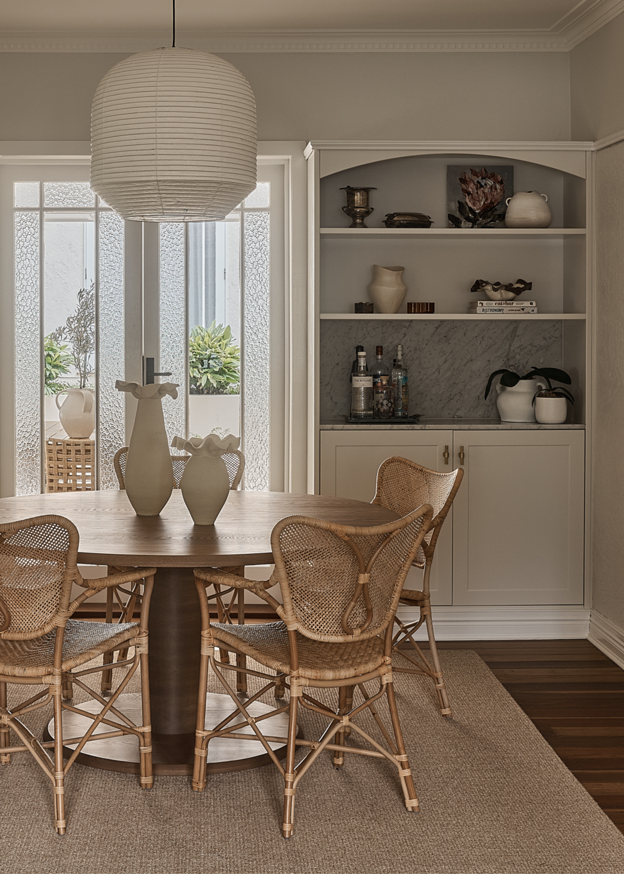A minimalist dining room with a wood table, rattan chairs, frilled vases, a paper pendant, and a nook with shelving and cabinets featuring bottles, vases, and potted plants