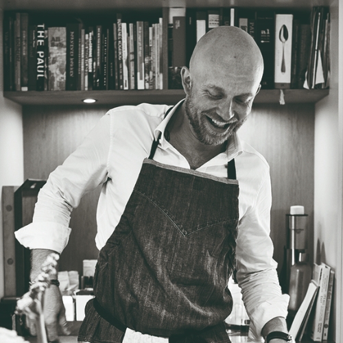 Black and white image of a man in a white button-down shirt with a dark apron on, standing in front of a desk with a shelf of books.