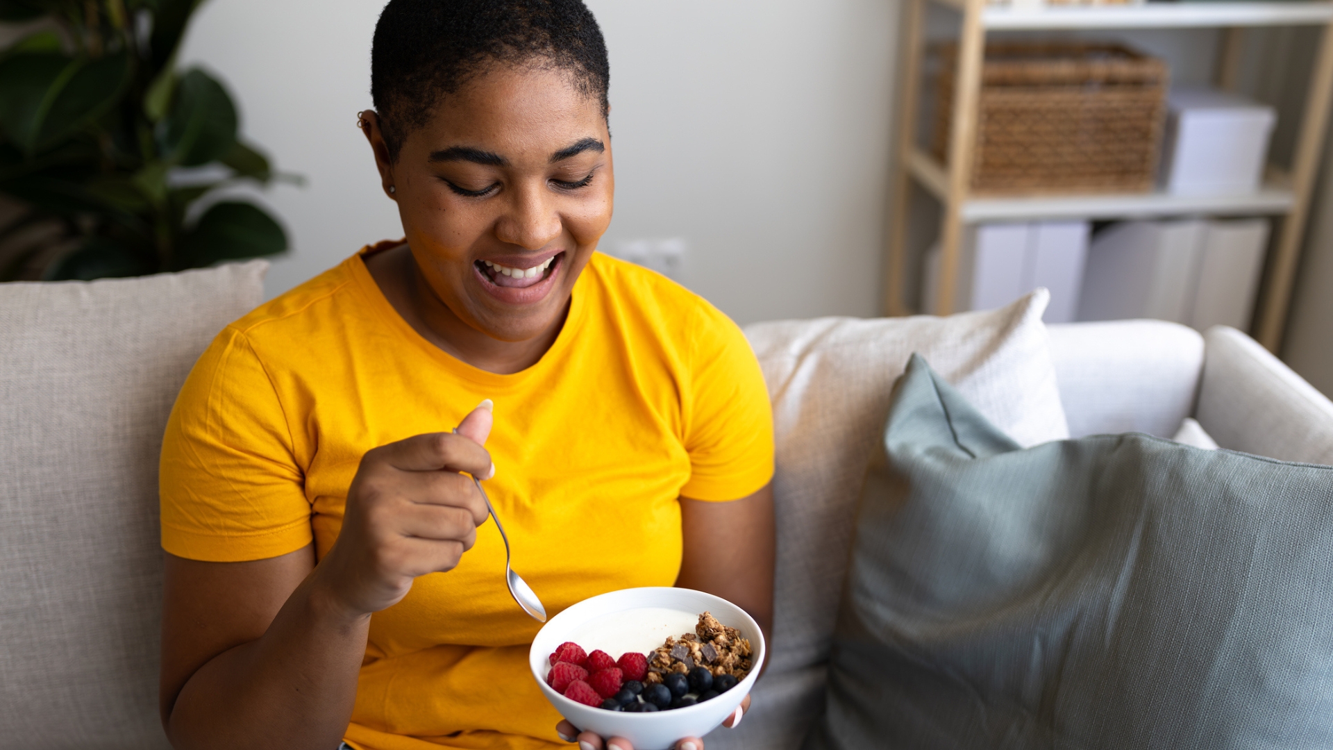 woman in a yellow tshirt sitting on a sofa close to holding a bowl of raspberries, blueberries and granola. shot from waist up and smiling at her bowl.