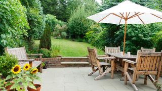 patio with wooden bench, table, chairs and parasol and steps leading up to a large lawn surrounded by trees and shrubs for privacy