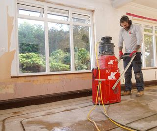 Man channelling out a concrete subfloor in a room with two windows, using the JK Floorheating system, ready for underfloor heating piping