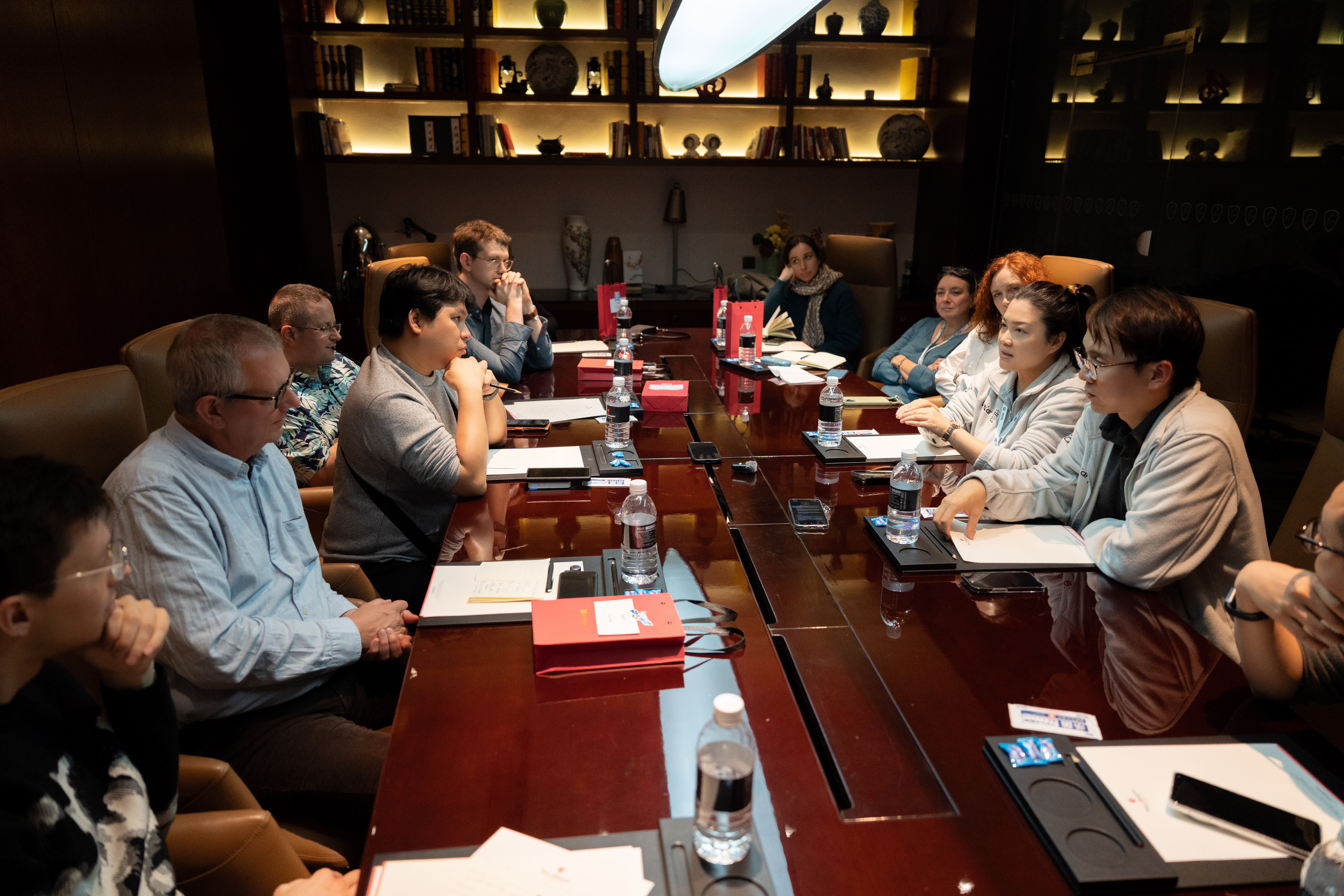A diverse group of professionals are seated around a long, polished wood conference table in a dimly lit meeting room, engaged in a collaborative discussion with notebooks and water bottles present.