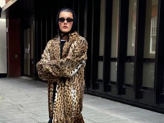 Fashion influencer @sobalera in London wearing a chic winter outfit with a knit triangle head scarf, a leopard print faux fur coat, oval sunglasses, black pants, and loafers.