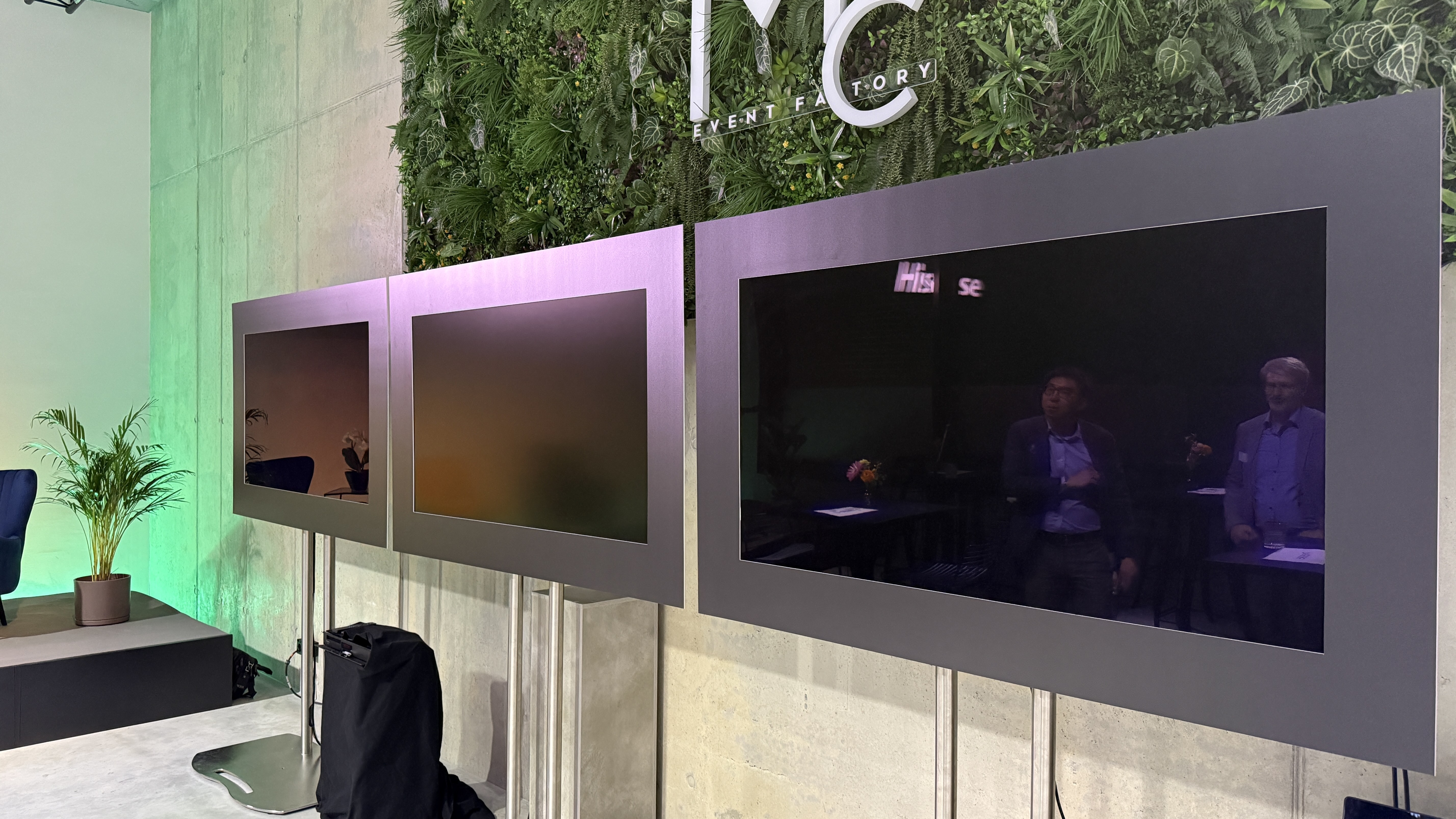 Three TVs are next to each other in an event space. They are the Samsung S90F, the Hisense UR9, and the LG C5. They are turned off, meaning you can see the reflection of what's around them, and the reflection on the Hisense is hazy, because of its antireflective screen