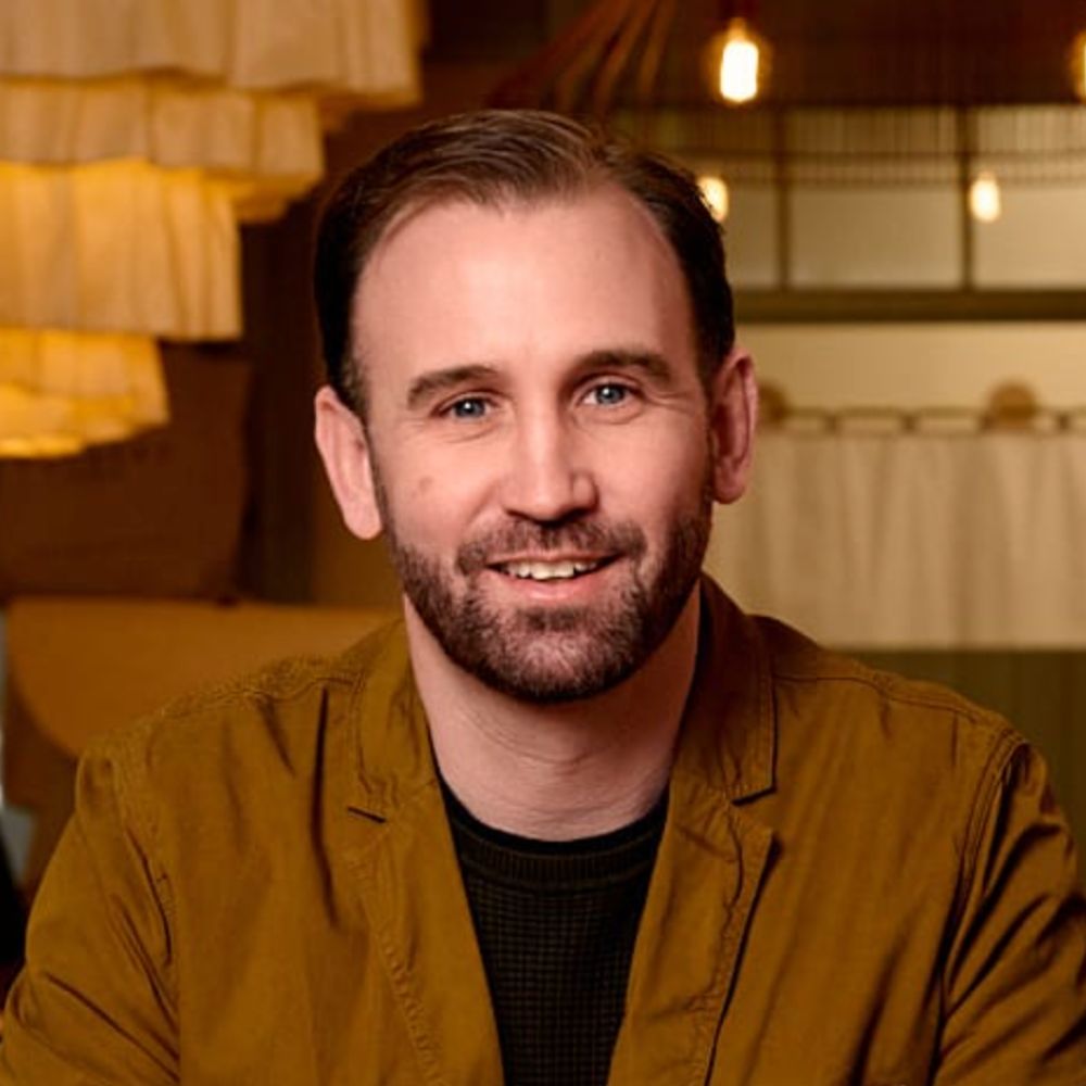 A head-and-shoulders portrait of a smiling man with short dark hair and a neatly trimmed beard. He wears a mustard-brown jacket over a dark shirt. Warm, amber lighting and softly glowing pendant lights create a cosy, blurred background behind him.