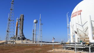 a tall orange rocket stands in the distance on the left, with a large white fuel tank in the foreground on the right.