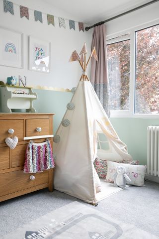 Kid's room with white teepee, oak chest of drawers, floral bunting, mint green wall paint to dado height and gold wall decals as scalloped border