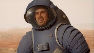 A very happy man wearing a blue astronaut suit and helmet. In the background you can see a red, rocky landscape.