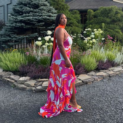 a photo of Mecca James-Williams in a pink, red, and white floral wedding guest dress standing in front of a house and flowers