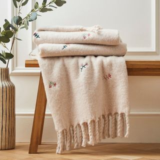 A cream throw with embroidered flowers on a wooden bench alongside a tall vase