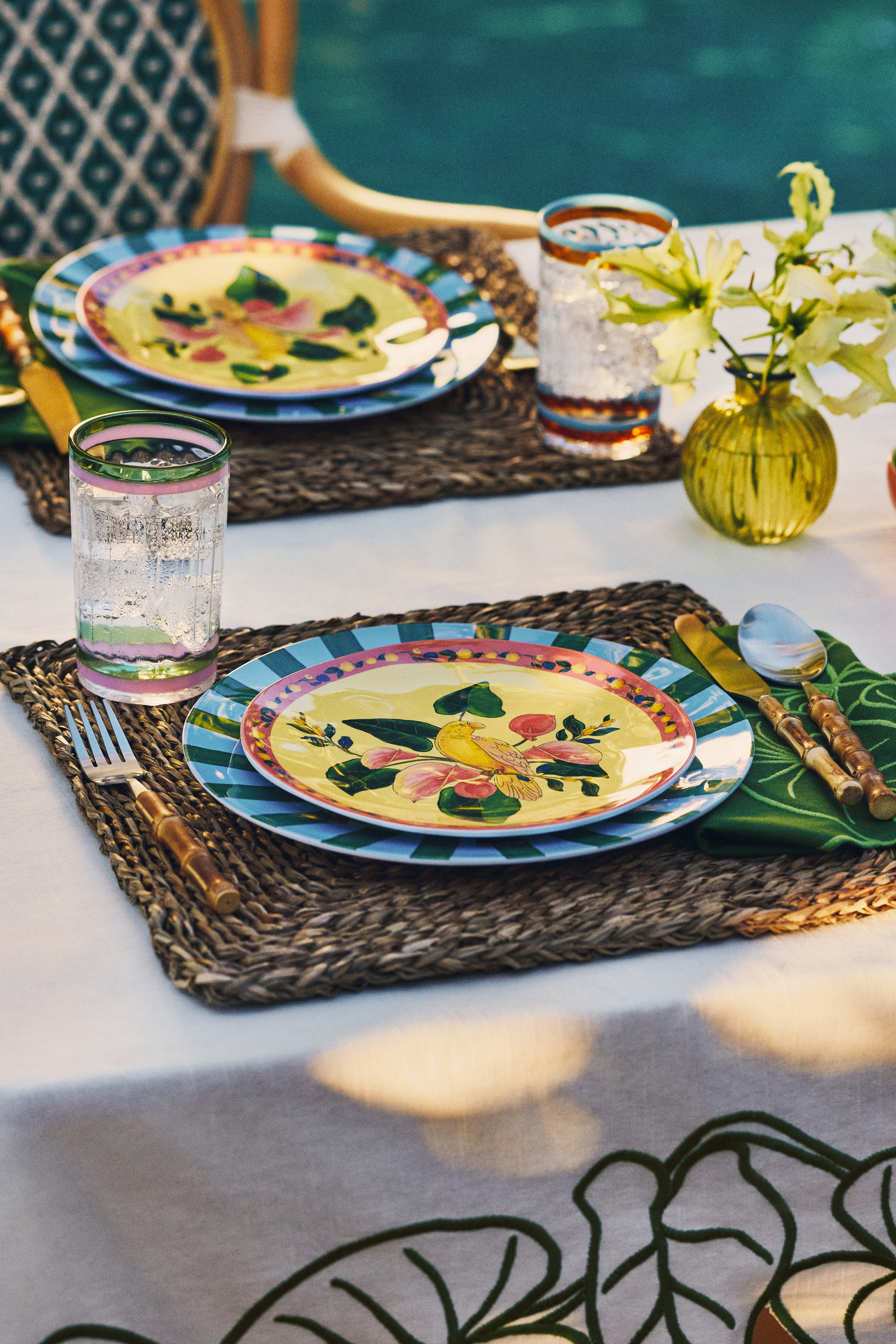 Image of two table settings on a white tablecloth. There are wicker placemats, blue and green striped chargers, yellow dinner plates, and bamboo cutlery.