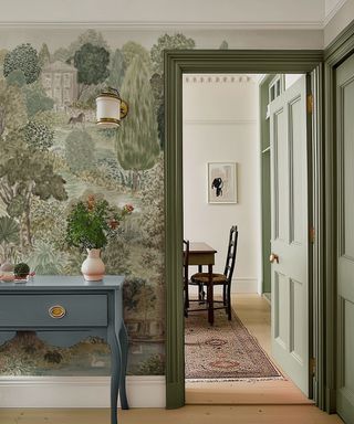 A hallway with a mural of a country garden scene. A pale blue console table with a vase of flowers sits against olive green woodwork and trim.