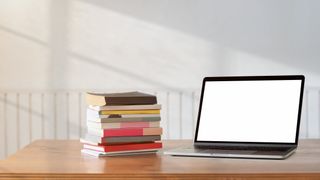 En laptop står öppnad på ett träfärgat skrivbord bredvid en stapel med böcker.