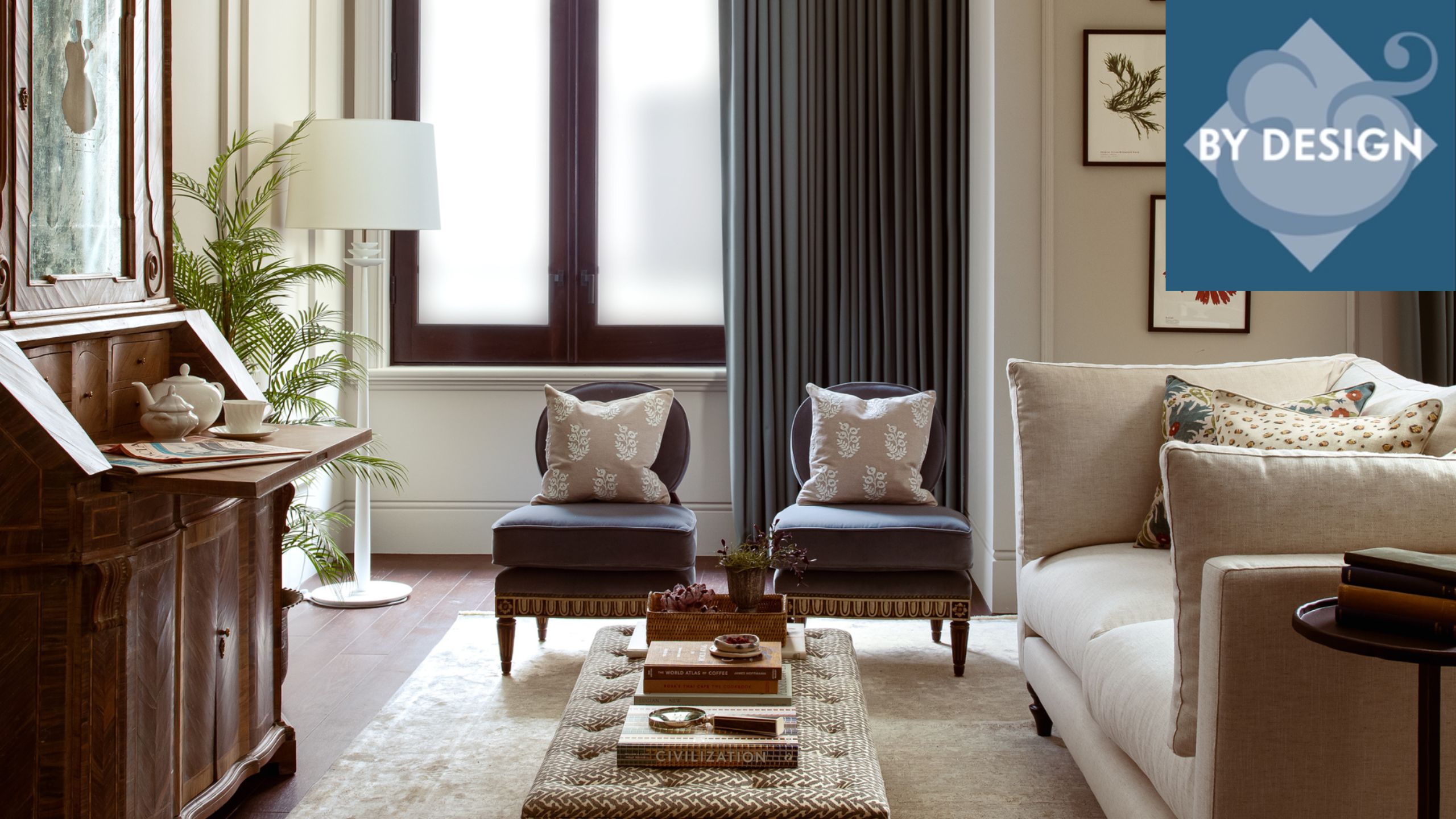 Neutral-toned living space with cream sofa, two navy armchairs positioned around upholstered ottoman, tall vintage wooden cabinet to left, windows in background with lamp and greenery