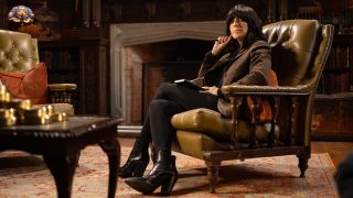 Host Claudia Winkleman sits by a fireside in a leatherbound chair in The Traitors U.K. season 2