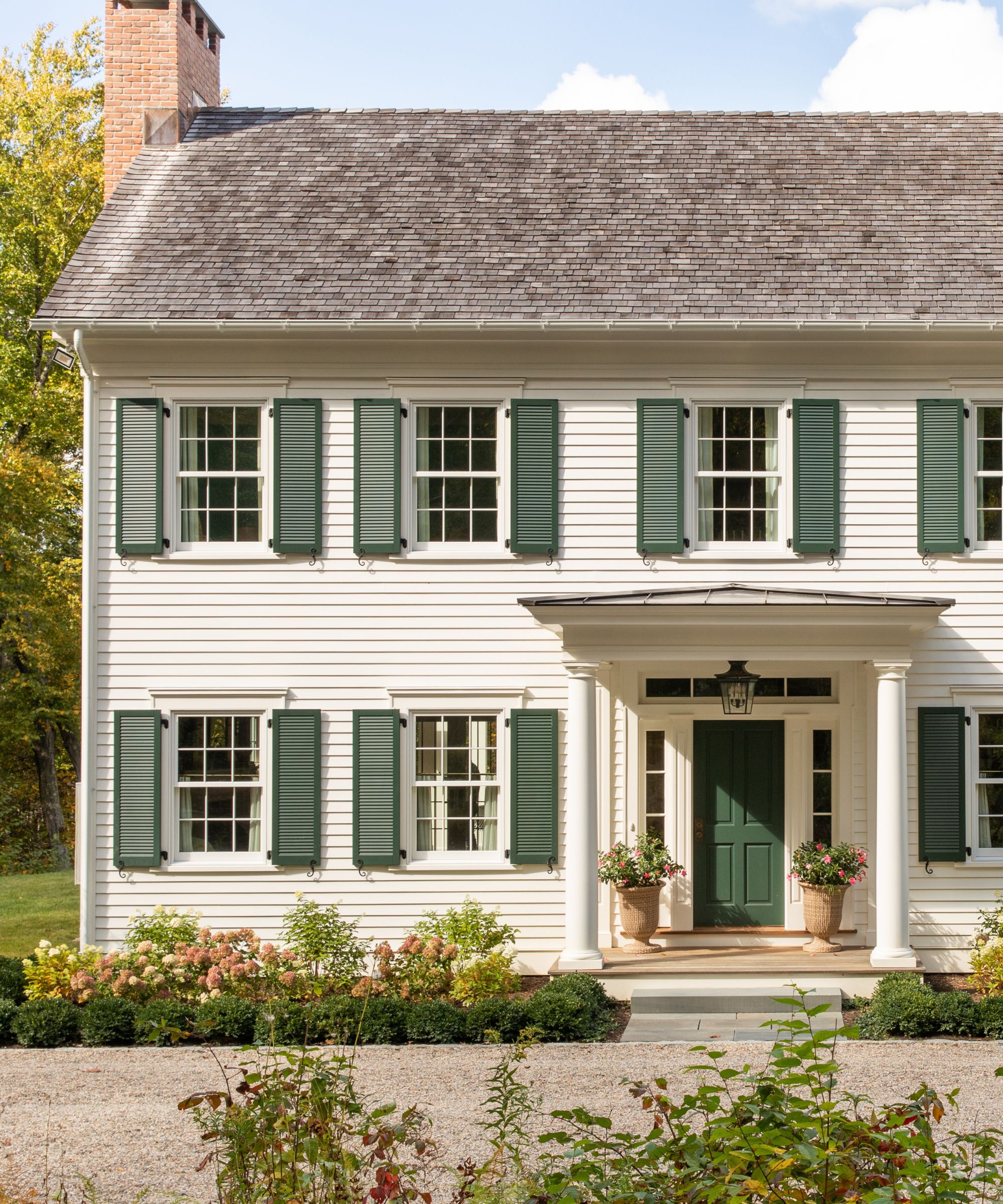 A traditional home exterior with warm white wood-clad walls, a slate roof, and dark green external shutters. A porch with a dark green front door and pots of flowers.