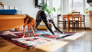 Woman stretching in living room on yoga mat with dog, surrounded by furniture and plants, after working out with sore muscles
