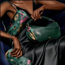A close-up of a female model wearing a black satin dress and holding three matching dark green and pink patterned hand bags.