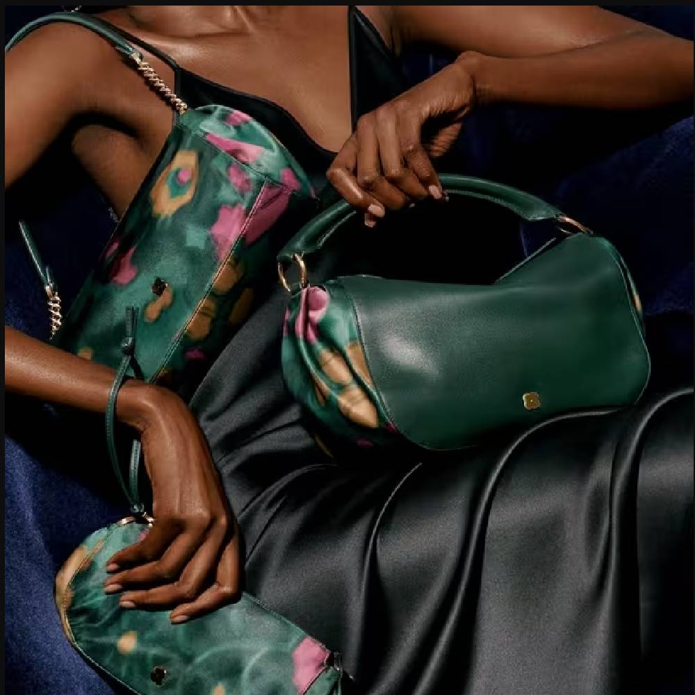A close-up of a female model wearing a black satin dress and holding three matching dark green and pink patterned hand bags.