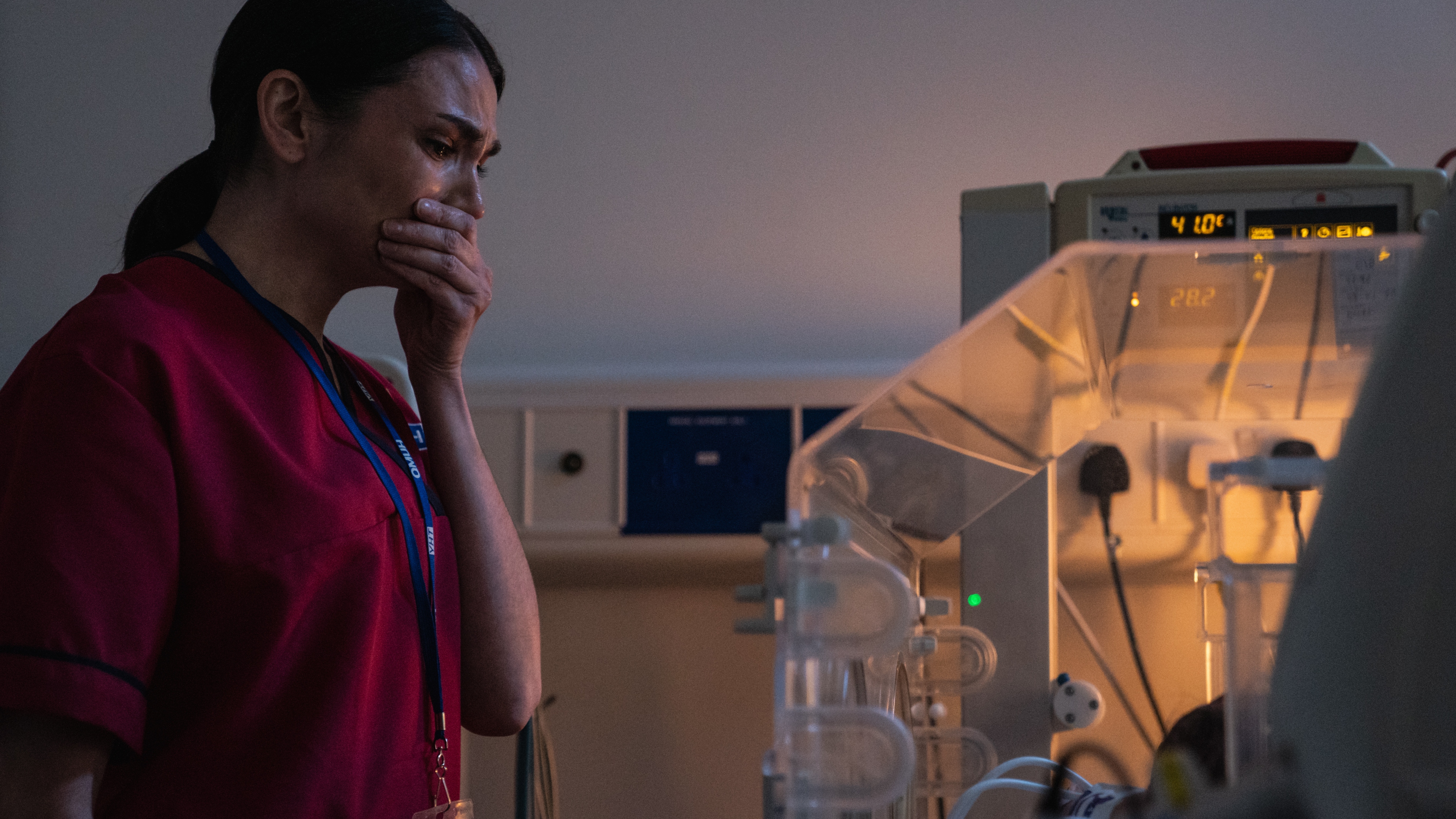 Sophia in her work scrubs crying with her hand to her mouth as she looks at a tiny baby in an incubator