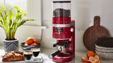 kitchenaid burr coffee grinder in red on a countertop with fruit, coffee, and croissants around it