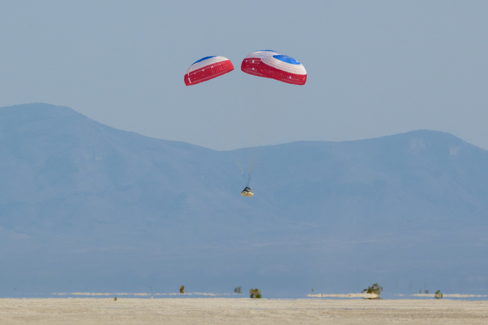 boeing's cst-100 starliner parachuting under three parachutes towards the desert with mountains in behind