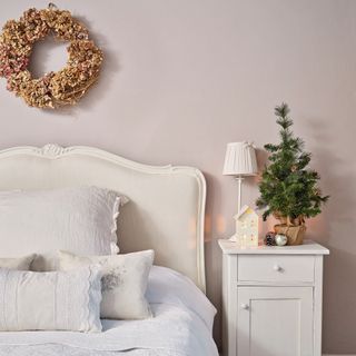 a bed with cream upholstered headboard, bedside table , mini Christmas tree and a wreath hung on the pale pink painted wall