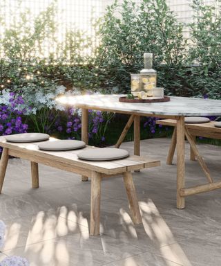 An outdoor living space with a marble table with lemon water on it, padded wooden benches either side of it, a concrete floor, and greenery around it