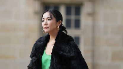 Yoyo Cao wears large golden earrings, a black faux fur fluffy long winter coat, a low-neck green dress, dark green tights, a green leather Givenchy bag, high heels shoes, outside Givenchy, during the Womenswear Spring/Summer 2024 as part of Paris Fashion Week on September 28, 2023 in Paris, France