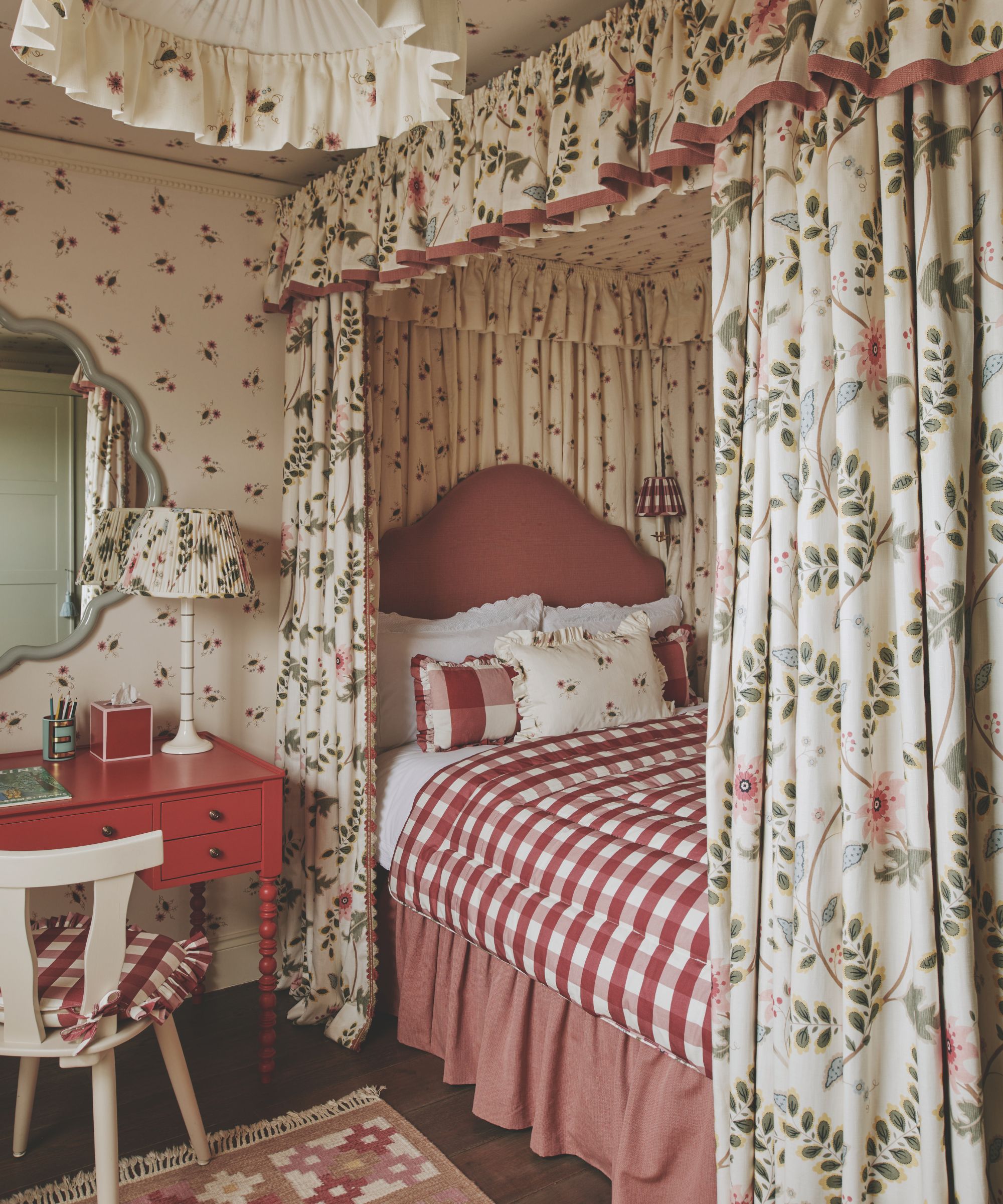 A single bed with a curtain surround and a red dressing table, lamp and chair.