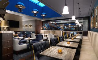 A restaurant in the Hilton London Bankside UK. White and brown marble tables, with deep blue, black, and light beige leather seating. There is a skyline on the ceiling with light fixtures hanging.