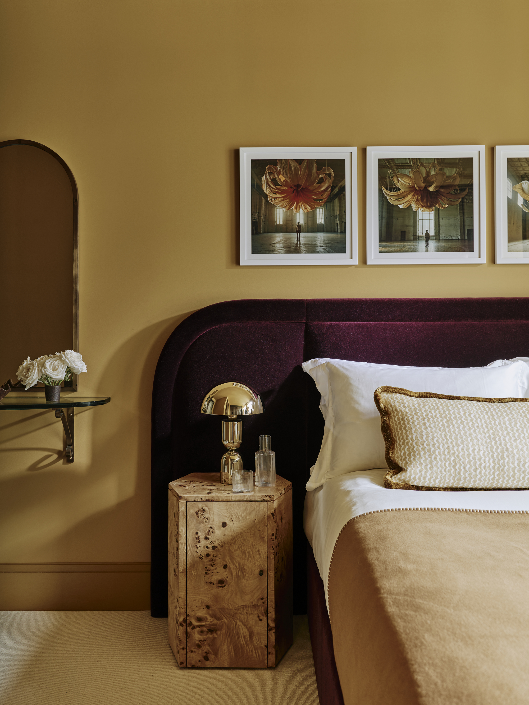 bedroom with yellow wall, burgundy headboard, burl side table, three artworks above the bed, white bedding with yellow blanket, and wall mirror